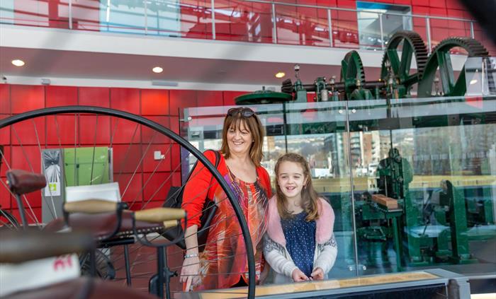 A mum stands with her young daughter inside the museum. They are looking at an early bicycle on display. The mum wears a red patterned dress and the g